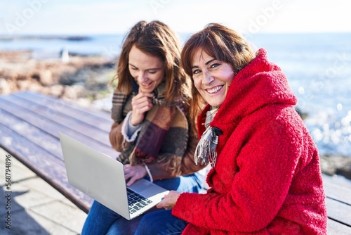 Two women mother and daughter using laptop sitting on bench at seaside