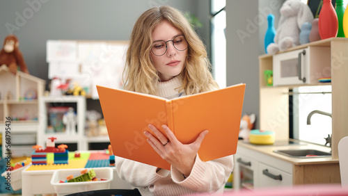 Young blonde woman preschool teacher reading book with relaxed expression at kindergarten