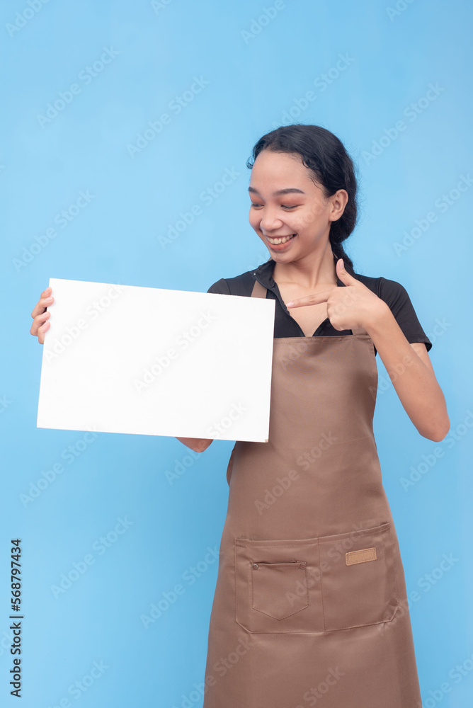 A young asian female waitress or barista points to a blank white signage she is holding. Wearing a brown apron and black shirt welcoming a customer and against a light blue background. A young asian female waitress or barista points to a blank white signage she is holding. Wearing a brown apron and black shirt welcoming a customer and against a light blue background.