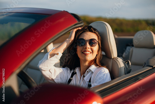 happy young beautiful woman in a white shirt sitting in a red car cabriolet with a white interior. red car in the field 
on a sunny day. the wind blows the girl's hair. red cabriolet. 