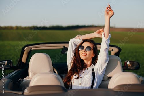 happy young beautiful woman in a white shirt sitting in a red car cabriolet with a white interior. red car in the field  on a sunny day. the wind blows the girl's hair. red cabriolet.  © Катя Іваськевич