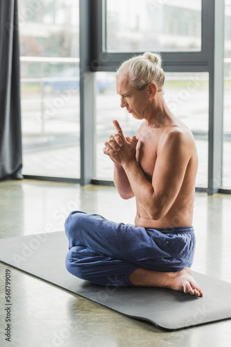 grey haired man in pants sitting in twisting yoga pose and doing crown chakra mudra.