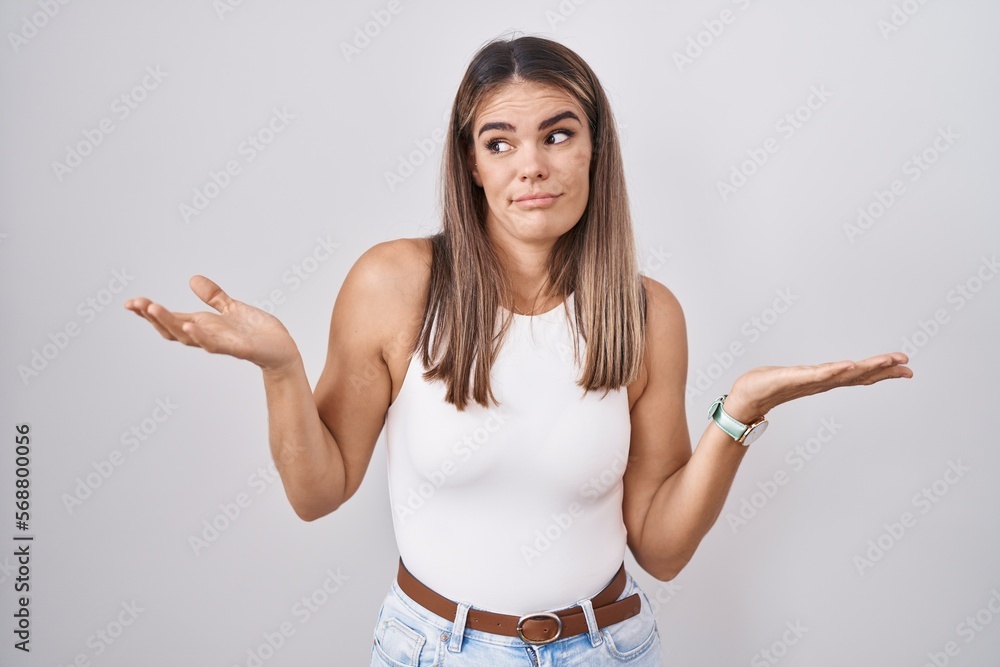 Hispanic young woman standing over white background clueless and confused expression with arms and hands raised. doubt concept. Hispanic young woman standing over white background clueless and confused expression with arms and hands raised. doubt concept.