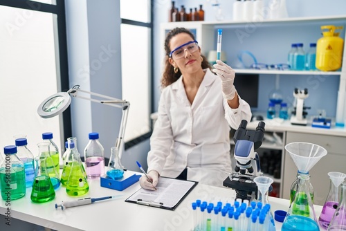 Young beautiful hispanic woman scientist writing on document holding test tube at laboratory