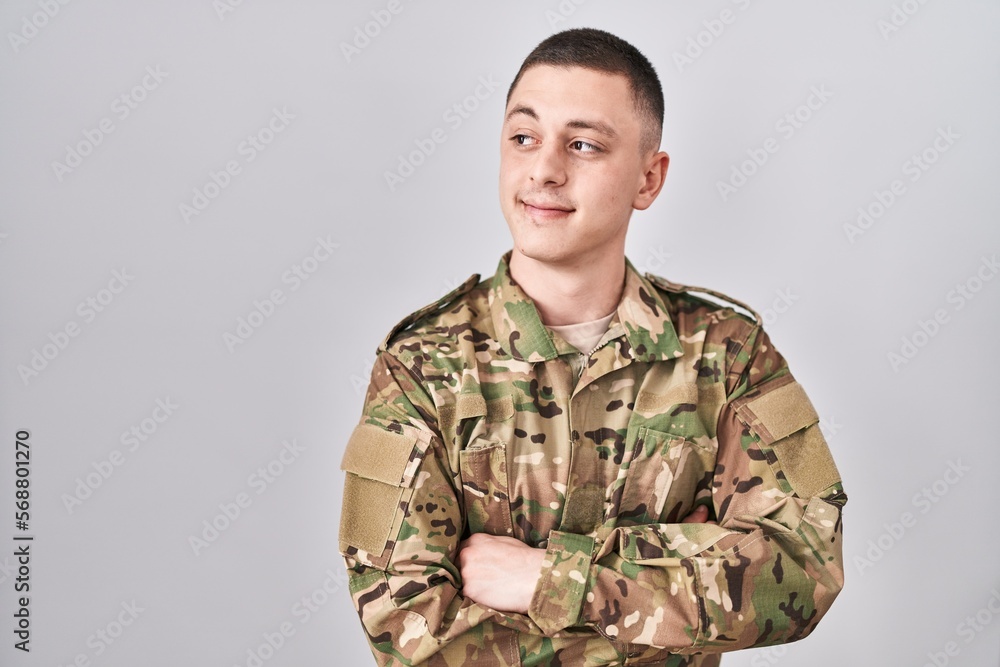 Young man wearing camouflage army uniform smiling looking to the side and staring away thinking.