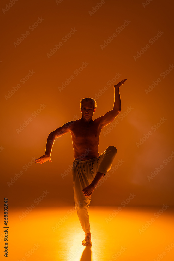 full length of shirtless man in pants practicing yoga on orange background.