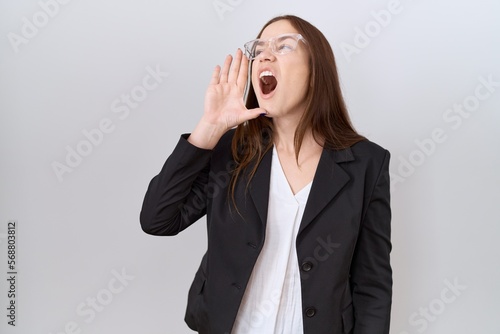 Beautiful brunette woman wearing business jacket and glasses shouting and screaming loud to side with hand on mouth. communication concept.