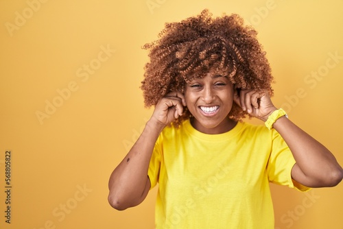 Young hispanic woman with curly hair standing over yellow background covering ears with fingers with annoyed expression for the noise of loud music. deaf concept.