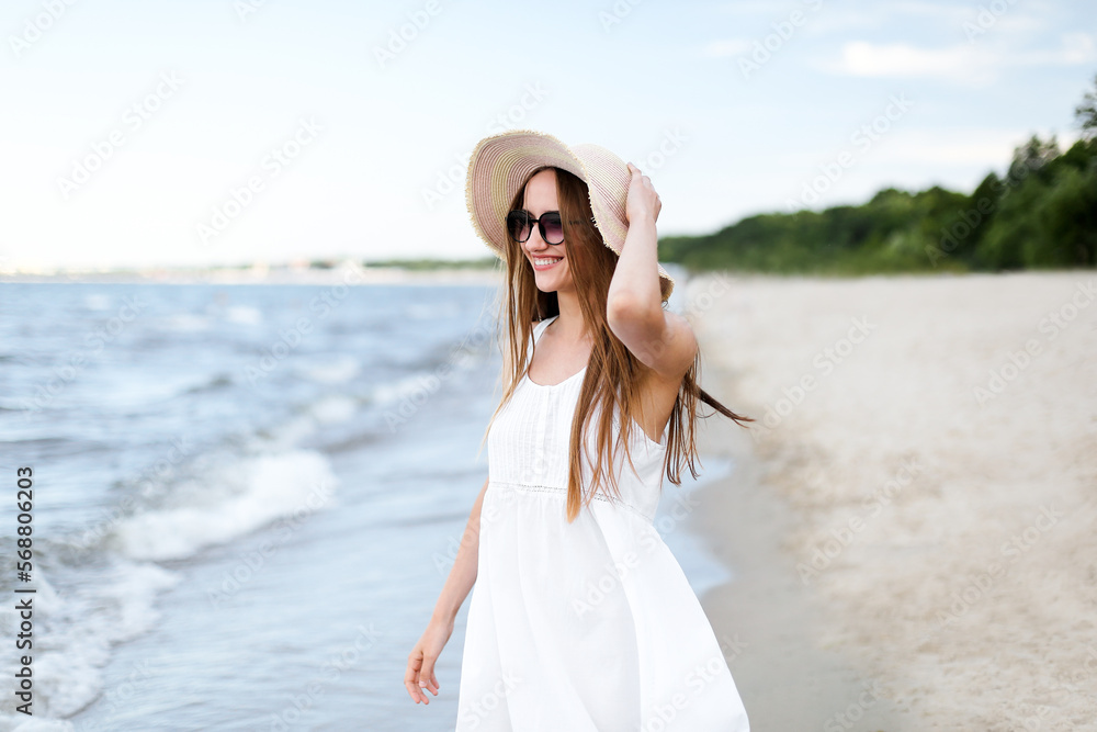 Happy smiling woman in free happiness bliss on ocean beach standing and posing with hat and sunglasses. Portrait of a female model in white summer dress enjoying nature Happy smiling woman in free happiness bliss on ocean beach standing and posing with hat and sunglasses. Portrait of a female model in white summer dress enjoying nature