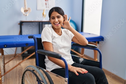 Young hispanic woman sitting on wheelchair at physiotherapy clinic smiling with hand over ear listening and hearing to rumor or gossip. deafness concept.