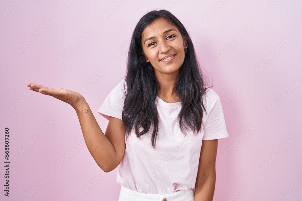 Young hispanic woman standing over pink background smiling cheerful presenting and pointing with palm of hand looking at the camera. Young hispanic woman standing over pink background smiling cheerful presenting and pointing with palm of hand looking at the camera.