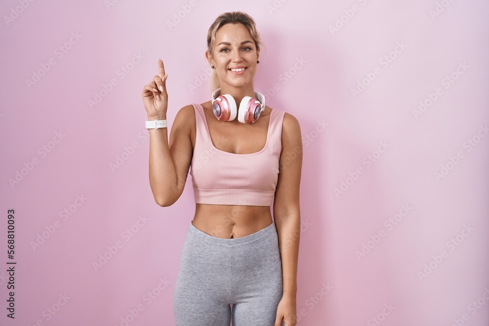 Young blonde woman wearing sportswear and headphones showing and pointing up with finger number one while smiling confident and happy.