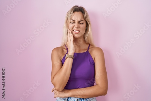 Young blonde woman standing over pink background touching mouth with hand with painful expression because of toothache or dental illness on teeth. dentist
