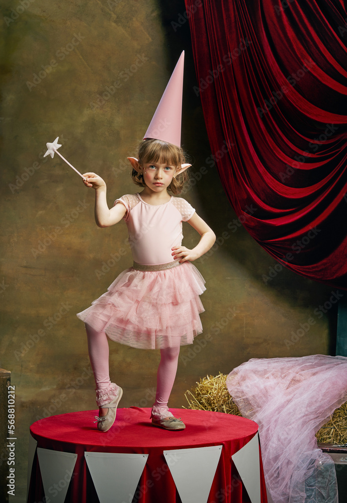Naklejka premium Magic. Little cute girl, kid in image of magical elf posing in pink dress over dark circus backstage background. Retro circus performance. Concept of dreams, art, fashion, vintage style.
