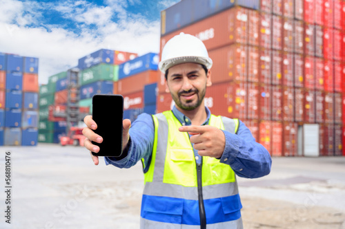 Logistic operators worker shows mock up mobile phone with blank screen, working in cargo container warehouse industry factory site in export, import, and transportation concept.