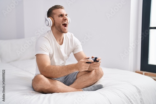 Young caucasian man sitting on the bed at home playing video games angry and mad screaming frustrated and furious, shouting with anger looking up.