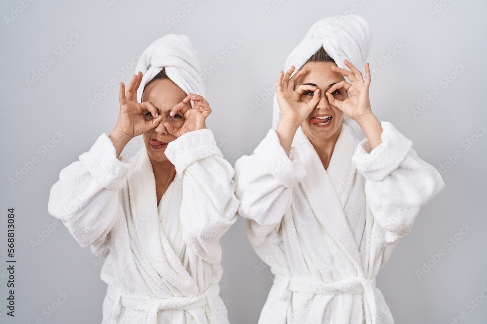 Middle age woman and daughter wearing white bathrobe and towel doing ok gesture like binoculars sticking tongue out, eyes looking through fingers. crazy expression. Middle age woman and daughter wearing white bathrobe and towel doing ok gesture like binoculars sticking tongue out, eyes looking through fingers. crazy expression.