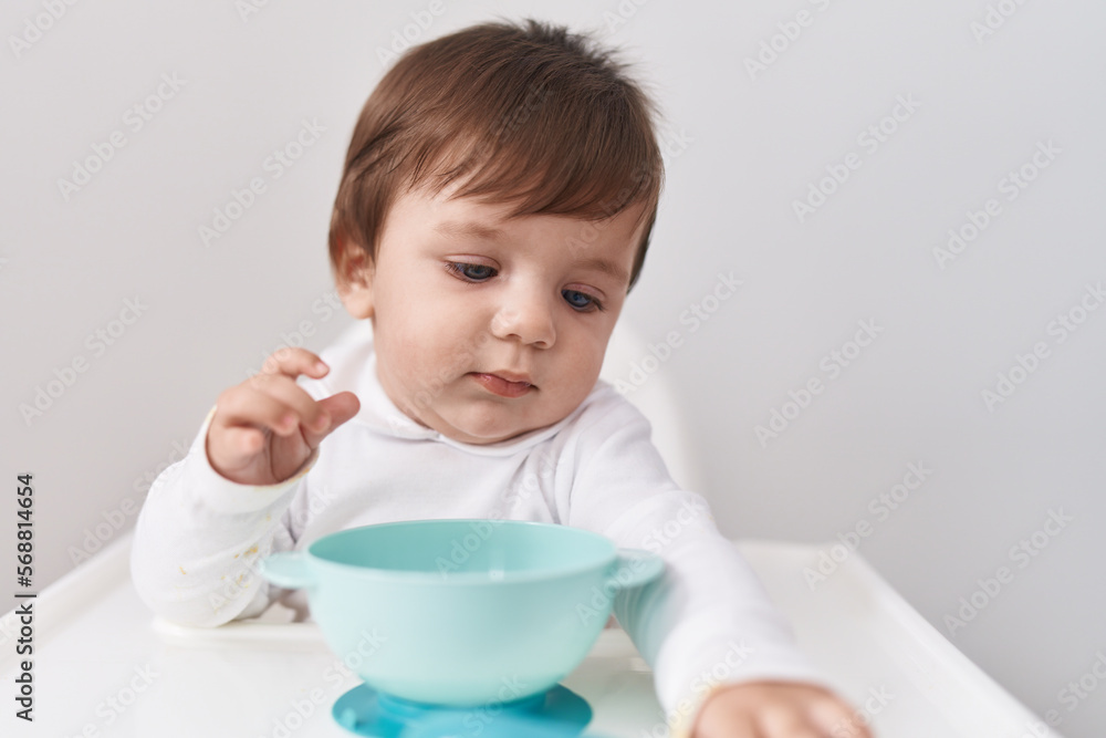 Adorable hispanic toddler sitting on highchair holding bowl over isolated white background Adorable hispanic toddler sitting on highchair holding bowl over isolated white background