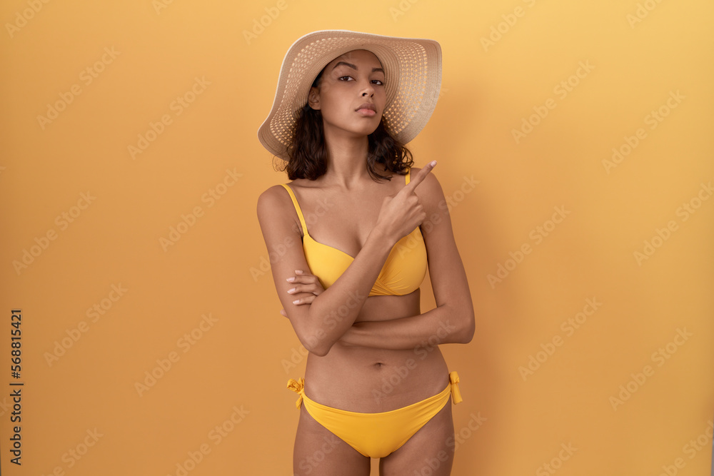 Young hispanic woman wearing bikini and summer hat pointing with hand finger to the side showing advertisement, serious and calm face Young hispanic woman wearing bikini and summer hat pointing with hand finger to the side showing advertisement, serious and calm face