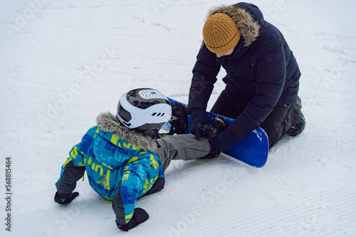 father helping little boy sitting on snow putting his feet in snowboard bindings adjusting straps.