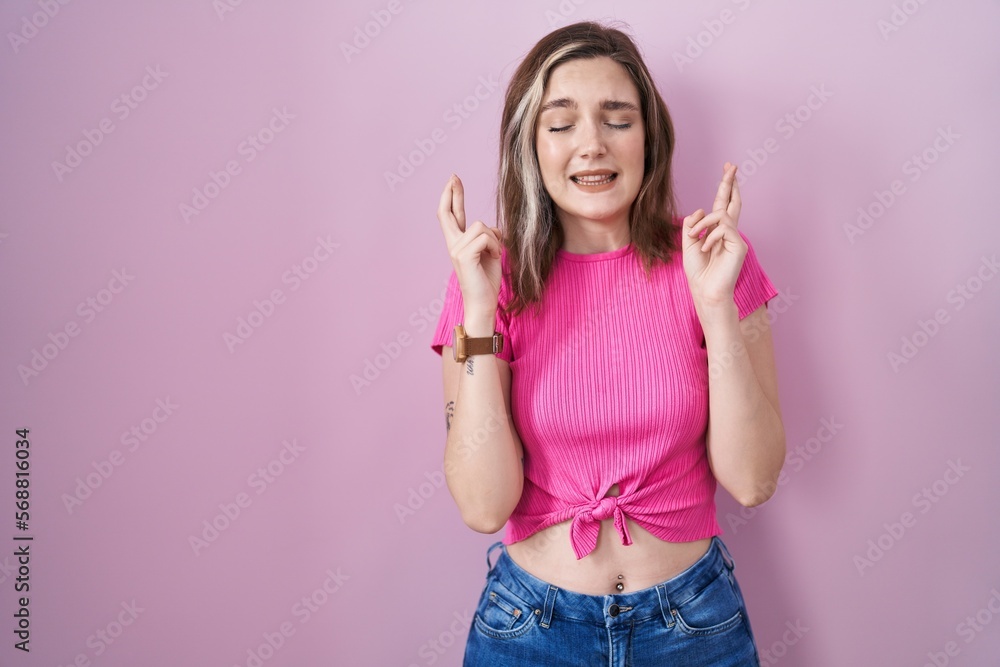 Blonde caucasian woman standing over pink background gesturing finger crossed smiling with hope and eyes closed. luck and superstitious concept. Blonde caucasian woman standing over pink background gesturing finger crossed smiling with hope and eyes closed. luck and superstitious concept.