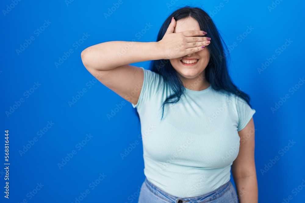 Young modern girl with blue hair standing over blue background smiling and laughing with hand on face covering eyes for surprise. blind concept. Young modern girl with blue hair standing over blue background smiling and laughing with hand on face covering eyes for surprise. blind concept.