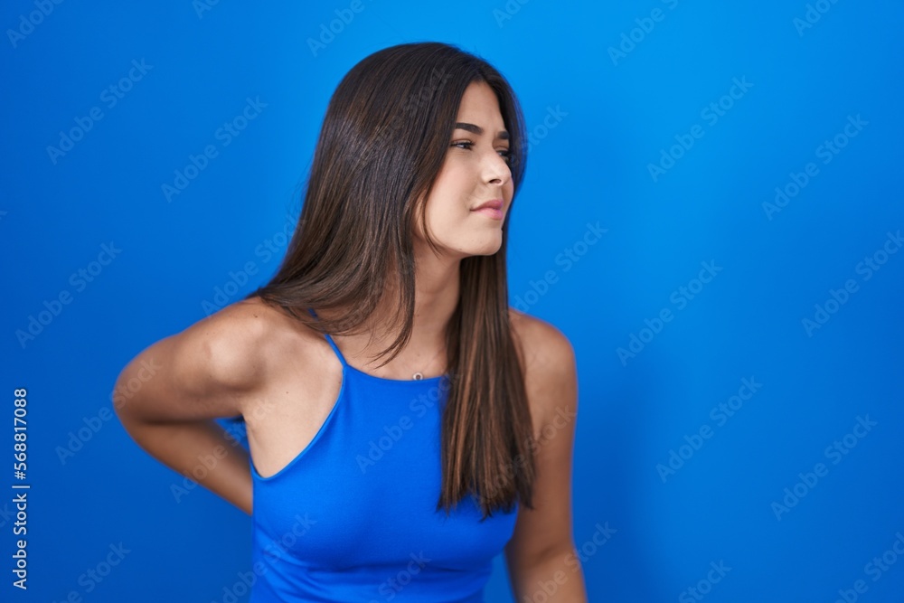 Hispanic woman standing over blue background suffering of backache, touching back with hand, muscular pain