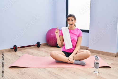Young brunette woman sitting on yoga mat at the gym pointing finger up with successful idea. exited and happy. number one.