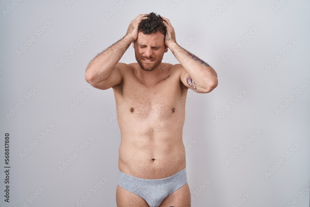 Young hispanic man standing shirtless wearing underware suffering from headache desperate and stressed because pain and migraine. hands on head.