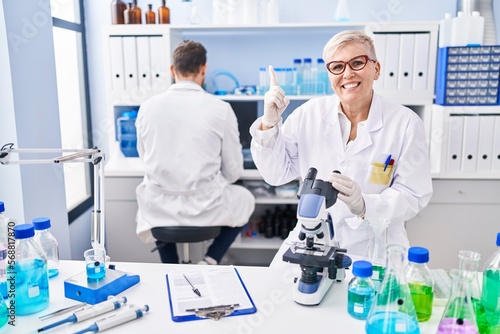 Middle age woman working at scientist laboratory smiling with an idea or question pointing finger with happy face, number one