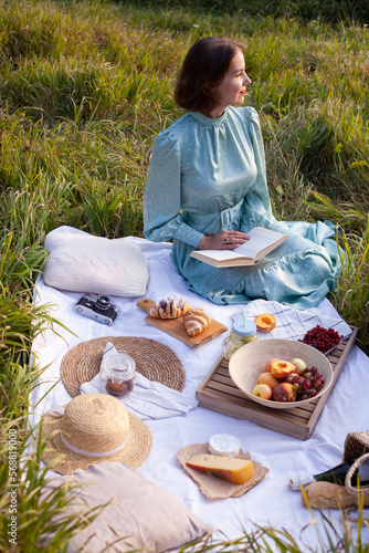 Wallpaper Mural A woman in a long summer dress with short hair sitting on a white blanket with fruits and pastries and reading the book. Concept of having picnic in a city park during summer holidays or weekends.  Torontodigital.ca