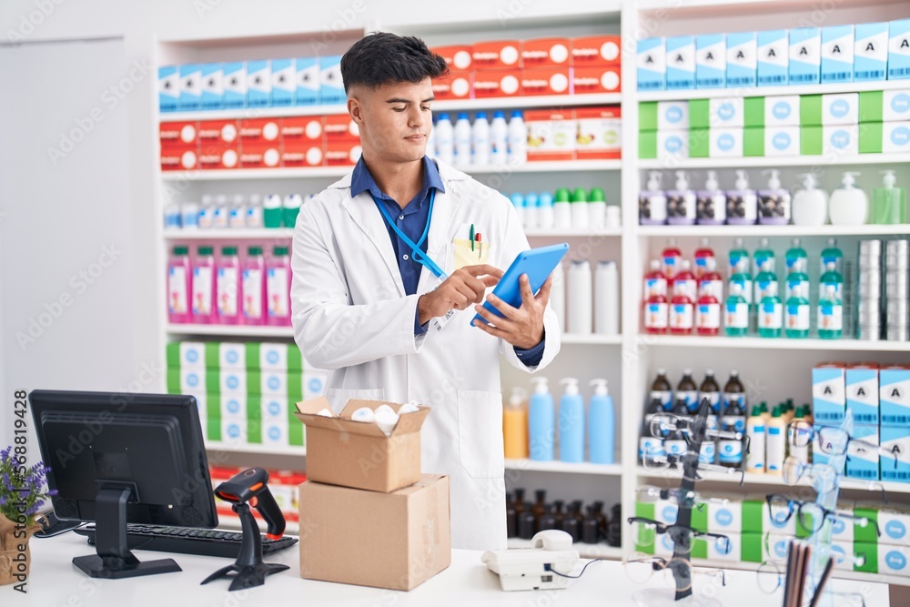 Young hispanic man pharmacist using touchpad holding pills bottle at pharmacy Young hispanic man pharmacist using touchpad holding pills bottle at pharmacy