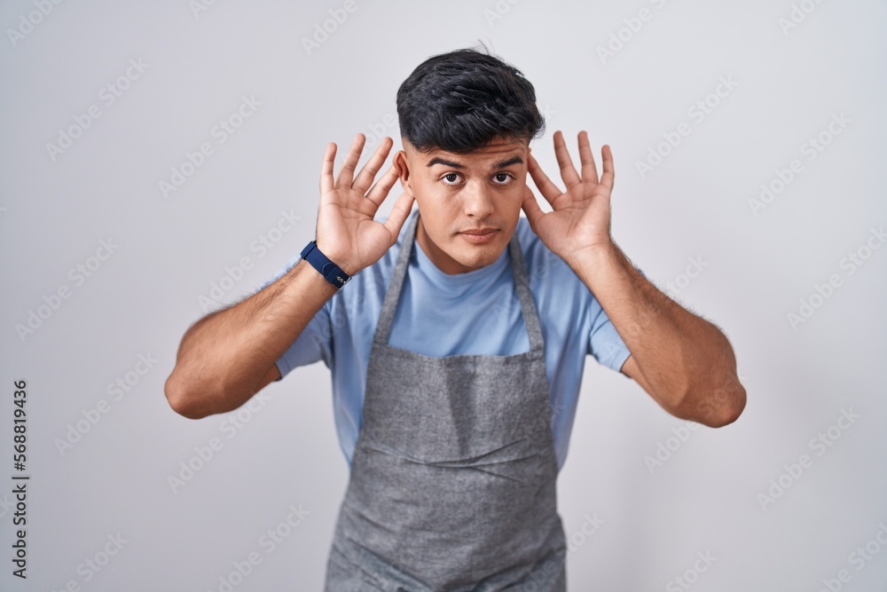 Hispanic young man wearing apron over white background trying to hear both hands on ear gesture, curious for gossip. hearing problem, deaf Hispanic young man wearing apron over white background trying to hear both hands on ear gesture, curious for gossip. hearing problem, deaf