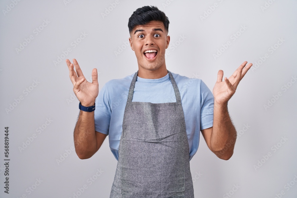 Hispanic young man wearing apron over white background celebrating crazy and amazed for success with arms raised and open eyes screaming excited. winner concept Hispanic young man wearing apron over white background celebrating crazy and amazed for success with arms raised and open eyes screaming excited. winner concept
