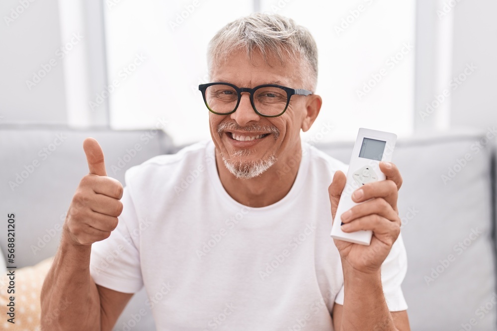 Hispanic man with grey hair holding air conditioner control smiling happy and positive, thumb up doing excellent and approval sign Hispanic man with grey hair holding air conditioner control smiling happy and positive, thumb up doing excellent and approval sign