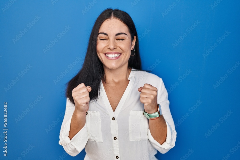 Young hispanic woman standing over blue background excited for success with arms raised and eyes closed celebrating victory smiling. winner concept. Young hispanic woman standing over blue background excited for success with arms raised and eyes closed celebrating victory smiling. winner concept.