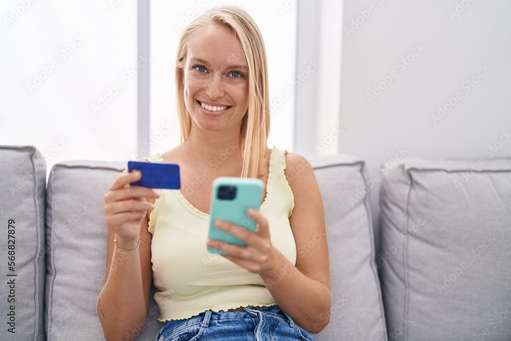 Young blonde woman using smartphone and credit card sitting on sofa at home Young blonde woman using smartphone and credit card sitting on sofa at home