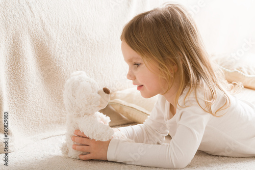 Bilde på lerret Adorable smiling little girl playing with white soft teddy bear on carpet at nursery room