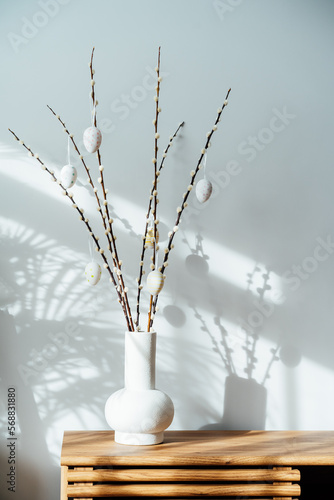 Easter home decor. Modern minimalist style interior with blooming branches of the pussy willow and hanging easter eggs in ceramic vase on a wooden console with sunlight and shadows on white gray wall.