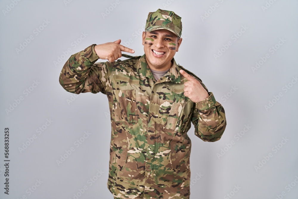 Hispanic young man wearing camouflage army uniform smiling cheerful showing and pointing with fingers teeth and mouth. dental health concept. Hispanic young man wearing camouflage army uniform smiling cheerful showing and pointing with fingers teeth and mouth. dental health concept.