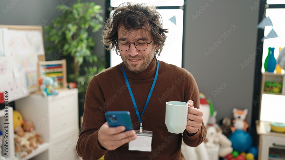 Young hispanic man preschool teacher using smartphone drinking coffee at kindergarten Young hispanic man preschool teacher using smartphone drinking coffee at kindergarten