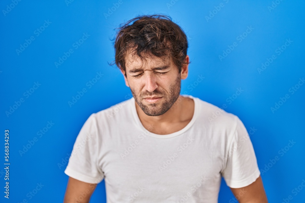 Hispanic young man standing over blue background with hand on stomach because indigestion, painful illness feeling unwell. ache concept. Hispanic young man standing over blue background with hand on stomach because indigestion, painful illness feeling unwell. ache concept.