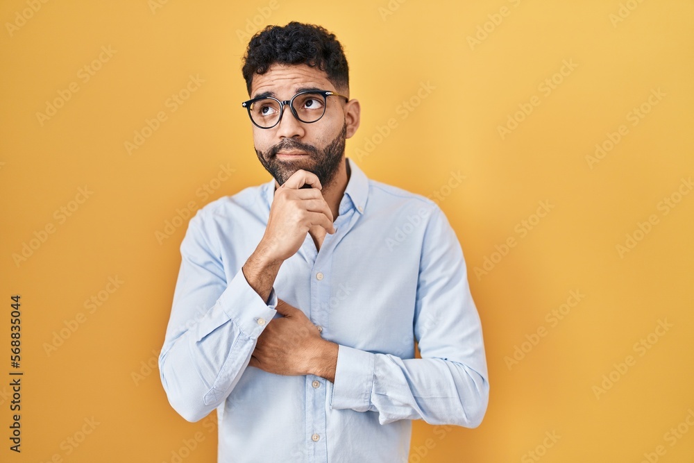 Hispanic man with beard standing over yellow background with hand on chin thinking about question, pensive expression. smiling with thoughtful face. doubt concept. Hispanic man with beard standing over yellow background with hand on chin thinking about question, pensive expression. smiling with thoughtful face. doubt concept.