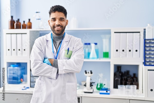 Young hispanic man wearing scientist uniform standing with arms crossed gesture at laboratory