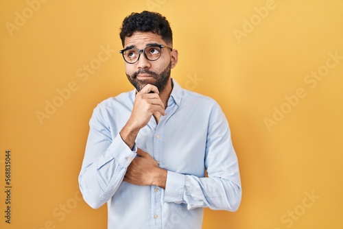 Hispanic man with beard standing over yellow background with hand on chin thinking about question, pensive expression. smiling with thoughtful face. doubt concept.