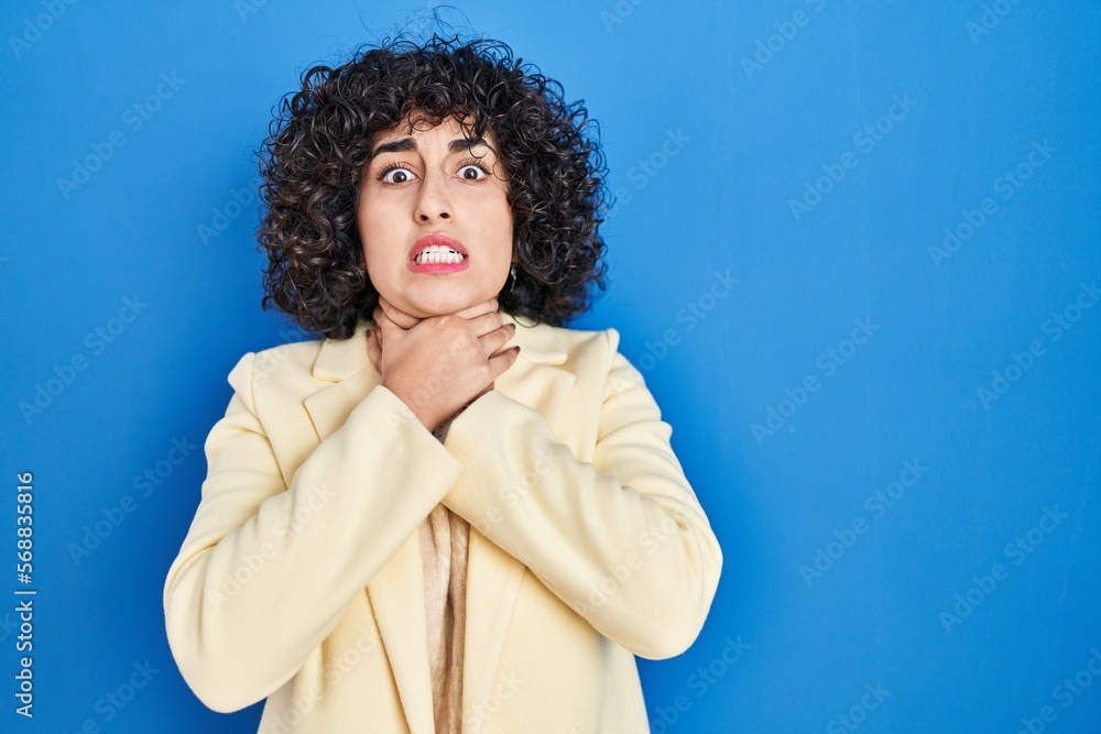 Young brunette woman with curly hair standing over blue background shouting suffocate because painful strangle. health problem. asphyxiate and suicide concept. Young brunette woman with curly hair standing over blue background shouting suffocate because painful strangle. health problem. asphyxiate and suicide concept.