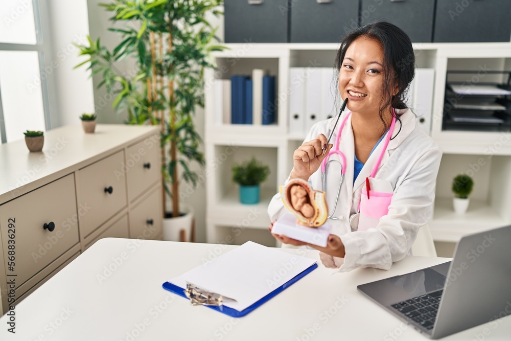 Young chinese woman wearing doctor uniform holding anatomical model of uterus with fetus at clinic Young chinese woman wearing doctor uniform holding anatomical model of uterus with fetus at clinic