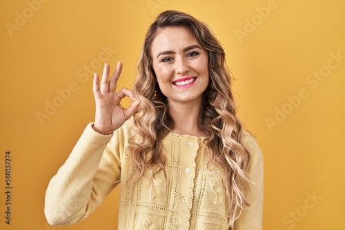 Young caucasian woman standing over yellow background smiling positive doing ok sign with hand and fingers. successful expression.