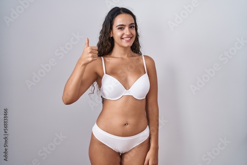 Young hispanic woman wearing white lingerie doing happy thumbs up gesture with hand. approving expression looking at the camera showing success.