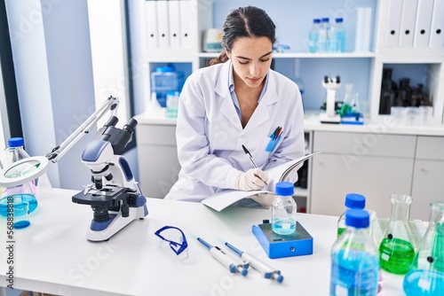 Young hispanic woman wearing scientist uniform measuring liquid at laboratory